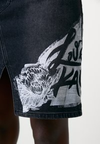 Črna denim krila z belim grafičnim tiskom cvetja in besedila. Oblikovanje vključuje ravni kroj in raztrgan rob.