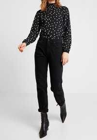Blusa a pois bianca e nera con collo alto e maniche arricciate, abbinata a jeans neri a vita alta e stivaletti neri a punta.