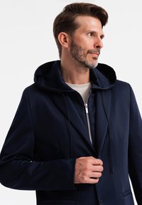 Marineblå zip-jakke med hætte, der har et glat stof, afslappet pasform og diskrete detaljer ved frontlommerne.