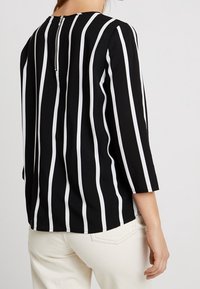 Kvinna i svart blus med vertikala vita ränder och blixtlås på övre delen av ryggen, ihopparad med gräddvitt byxor.