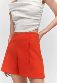 Kvinde iført klare orange højtaljede skræddersyede shorts og en ærmeløs hvid rynket crop top, stående med den ene hånd på hoften.