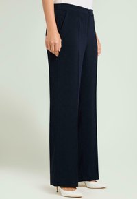 Marineblauwe wijde broek van een gladde stof; voorzien van voorplooien en zakken aan de zijkant; gecombineerd met witte hakken.