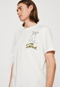 Camisa de algodón blanca con un contorno negro de un personaje con los brazos levantados y el logo de "Los Simpson" en amarillo. Mangas cortas, ajuste holgado.