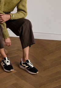 Schwarze und cremefarbene Sneakers aus Wildleder und Mesh-Materialien, mit einem kontrastierenden Leopardenmuster an der Rückseite und weißen Schnürsenkeln, kombiniert mit braunen Cropped-Hosen.