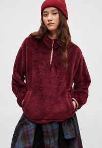 Bordowy polarowy pulower z wysokim kołnierzem i zamkiem do ćwiartki, z bocznymi kieszeniami i miękką, teksturowaną powierzchnią. Warstwowo noszony z koszulą w kratę.