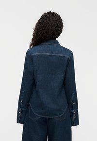 Σκούρο πουκάμισο από τζιν με γιακά, μανσέτες με κουμπιά και καμπυλωτό τελείωμα. Διαθέτει ραφές και δύο πίσω τσέπες.