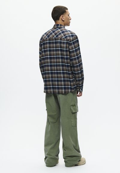 Chemise à carreaux dans des teintes marine, marron et bleu clair avec un col boutonné ; associée à un pantalon cargo vert foncé et des chaussures beige.