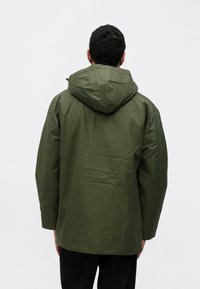 Casaco impermeável verde com capuz, apresentando uma textura lisa e corte recto. A parte de trás não tem marcas visíveis ou adereços.