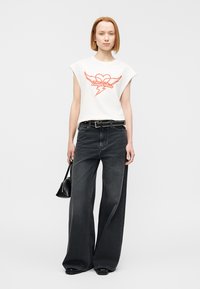 Λευκό αμάνικο μπλουζάκι με κόκκινο γραφικό, σε συνδυασμό με μαύρο τζιν ψηλόμεσο παντελόνι με φαρδιά πόδια. Συμπεριλαμβάνονται μαύρη ζώνη και μικρή μαύρη τσάντα.