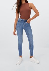 Jeans de mezclilla azul ajustados con una cintura alta, que presentan un diseño de cinco bolsillos, un ligero desgastado y una textura suave, combinados con una blusa sin mangas marrón.