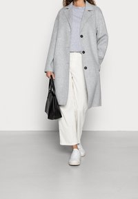 Grå ylleblandad kappa med avslappnad passform, svarta knappar, vita texturerade vida byxor och vita sneakers. Svart handväska i handen.