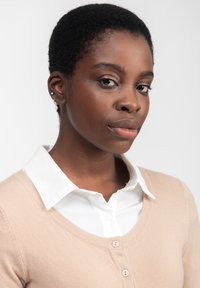 Beige Cardigan über einem weißen Hemd mit Kragen, das einen V-Ausschnitt und drei rosa Knöpfe hat. Kurze, strukturierte schwarze Haare und elegante Ohrringe sind sichtbar.