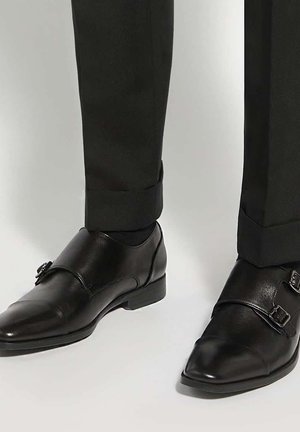 Zwarte leren monkstrap herenschoenen gedragen met een zwarte broek tegen een effen lichte achtergrond.