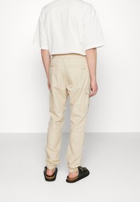 Pantaloni cargo beige con polsini elasticizzati, multiple tasche laterali e una vestibilità rilassata. Indossati con sandali neri scivolati e una t-shirt bianca ampia.