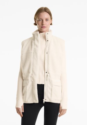 Nő krémszínű fleece mellényt visel egy világos cipzáras felső fölött és fekete leggingset, fehér háttér előtt áll.
