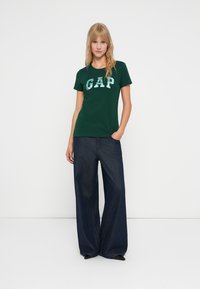 Camiseta de algodón verde con el logo "GAP" en azul claro, combinada con vaqueros de pierna ancha de denim oscuro. El modelo está de pie con una mano en el bolsillo.