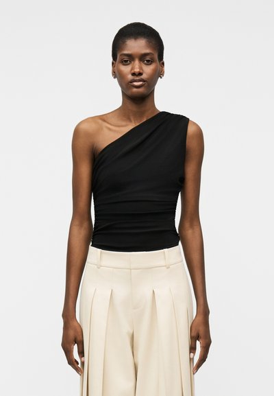 Femme portant un haut noir à une épaule et un pantalon crème plissé taille haute, debout devant un fond clair uni.