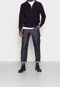 Pull en maille bleu marine avec une fermeture éclair au quart, superposé à un T-shirt blanc, associé à un jean en denim foncé retroussé aux poignets et des bottines noires.