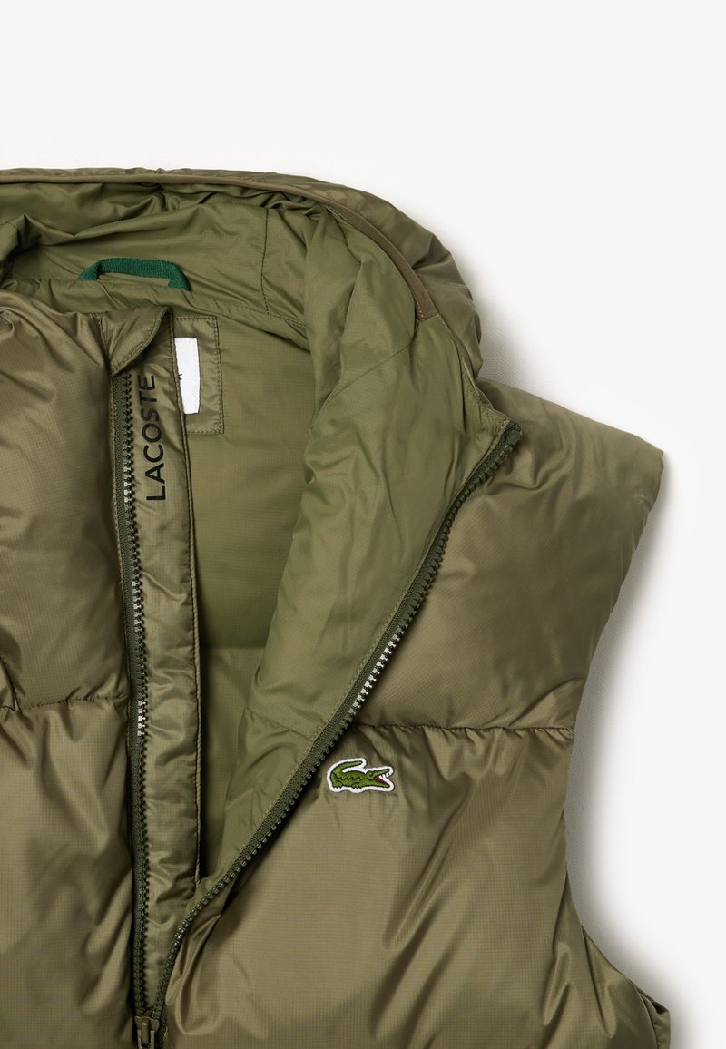 Chaleco acolchado verde oliva con cierre frontal, interior forrado y acento del logo de Lacoste. Textura suave con un acabado ligeramente brillante.