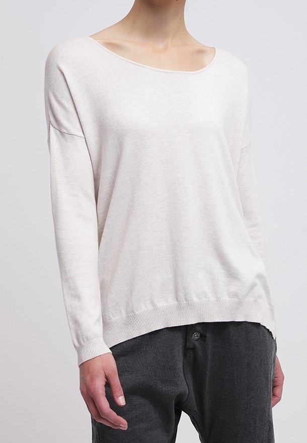 Personne portant un pull ample à manches longues de couleur beige clair avec un large col et un pantalon gris foncé sur un fond uni.