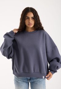 Üks suur, ümarkaelusega dressipluus pehmes tumedas hallis kangas, langenud õlgade ja soonikservadega, koos helesiniste teksadega.