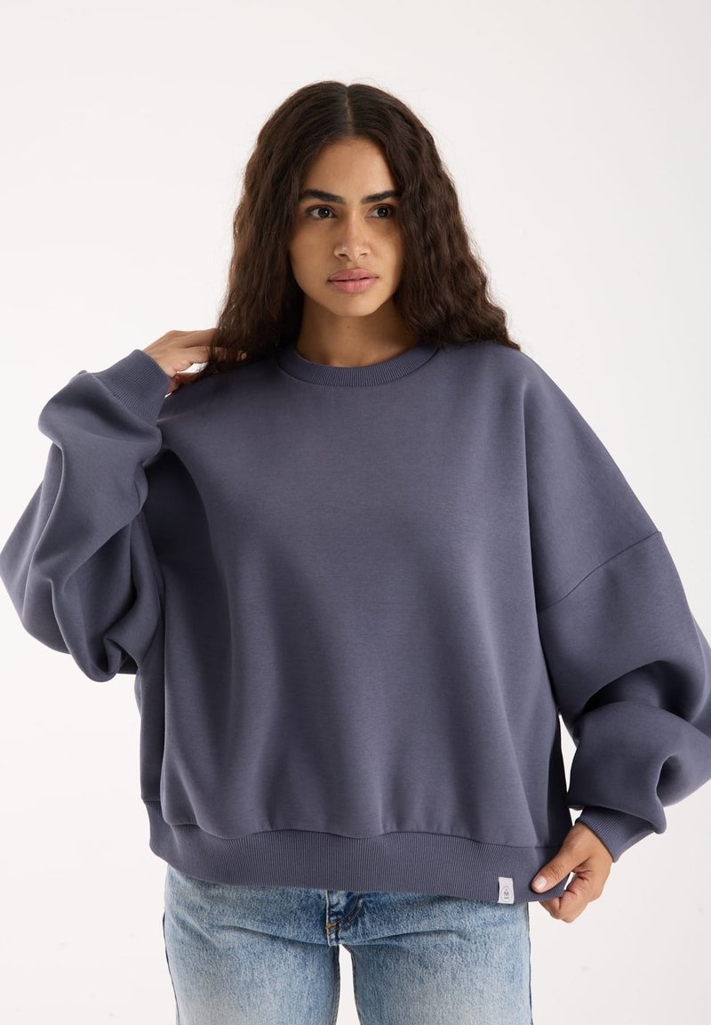 Üks suur, ümarkaelusega dressipluus pehmes tumedas hallis kangas, langenud õlgade ja soonikservadega, koos helesiniste teksadega.