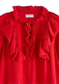 Rød bluse med flæsekrave og detaljer foran, lavet af letvægtsstof. Har en knyttet lukning ved halsen og en tekstureret overflade.