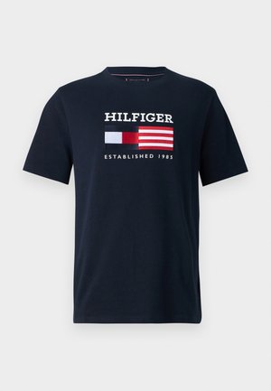 Marineblaues T-Shirt mit kurzen Ärmeln, dem Schriftzug „HILFIGER“, einem roten, weißen und schwarzen rechteckigen Logo und dem Schriftzug „ESTABLISHED 1985“ darunter.