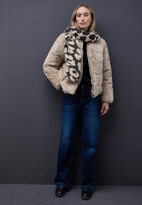Veste en fausse fourrure beige clair avec fermeture éclair à l'avant, associée à un jean large bleu foncé. Une écharpe à imprimé animalier ajoute un contraste de motifs. Des chaussures noires complètent le look.