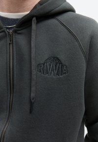 Sweat à capuche gris foncé zippé avec un logo devant indiquant "RIVVIA" en broderie noire en relief et des cordons à la capuche.