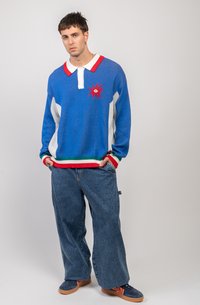 Pulover tricotat albastru cu guler roșu, accente albe și un grafic cu soare; pantaloni din denim lejeri cu buzunare mari; pantofi slip-on maro.