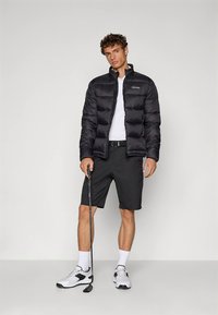 Svart vadderad jacka med hög krage och lätt glans, bärs över en vit pikétröja, kombinerad med svarta shorts och vita sportskor.