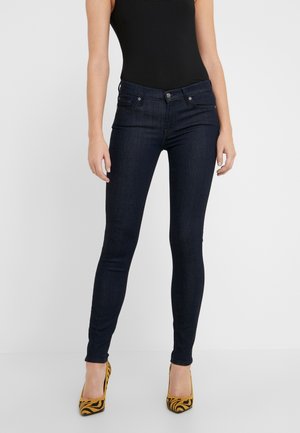 Femme portant un jean skinny foncé, un haut noir sans manches et des talons hauts jaunes à imprimé animal, debout devant un fond uni.