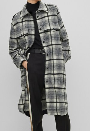 Frau trägt einen langen schwarz-weiß karierten Mantel über einem schwarzen Rollkragenpullover und schwarzen Hosen mit weißem Seitenstreifen.