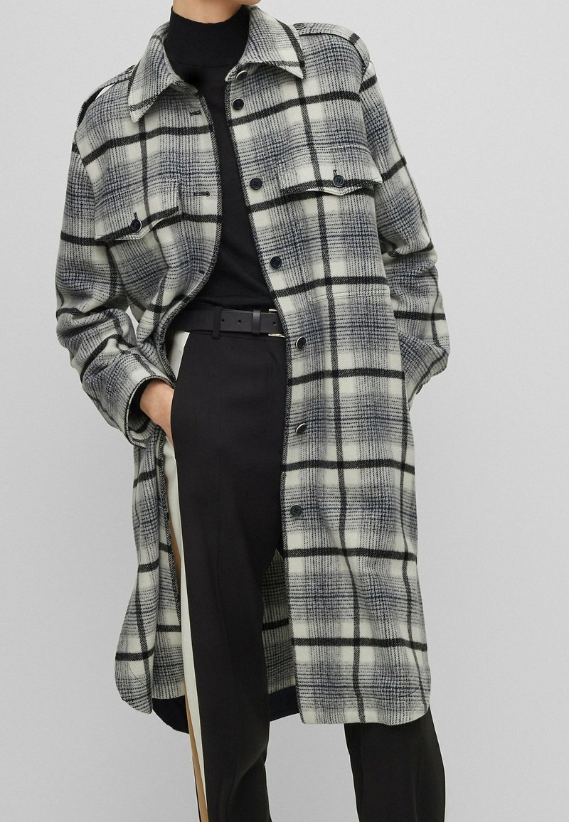 Femme portant un long manteau à carreaux noir et blanc par-dessus un col roulé noir avec un pantalon noir présentant une bande blanche sur le côté.