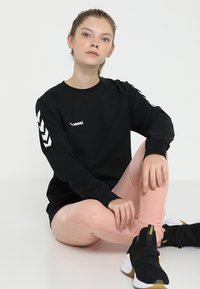 Jonge vrouw zit met gekruiste benen, draagt een zwarte Hummel trui, roze legging en zwarte Puma sneakers tegen een effen witte achtergrond.