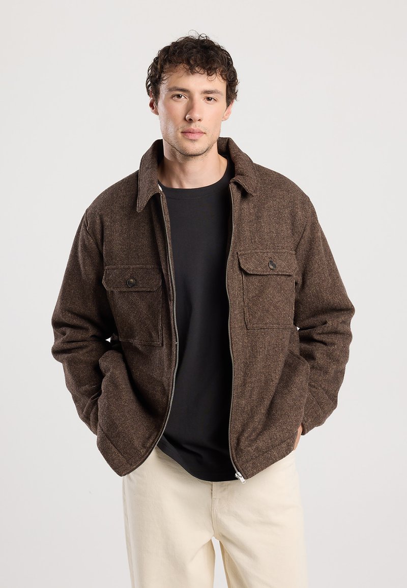 Brown, textured jacket with a zip front, two chest pockets, and a collared neck. Worn over a black shirt, with minimalistic design.
