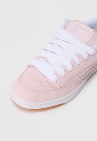 Svijetloružičasta suede tenisica s bijelim vezicama, koja ima perforirani prednji dio, gumeni potplat i mali Reebok logo na jeziku.