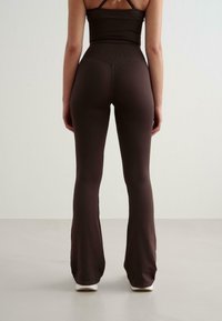 Femme debout contre un fond uni portant un pantalon ample marron foncé et un crop top près du corps assorti, vue de dos.