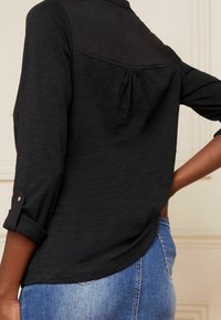 Musta pitkäripsinen paita takana olevalla kerätty yksityiskohdalla, jossa on sileä pinta ja käänteet, yhdistettynä vaaleansinisiin farkkuihin.