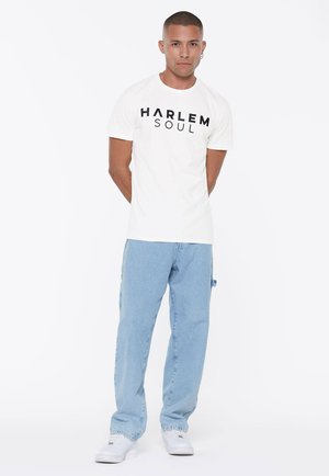 Weißes Baumwoll-T-Shirt mit den in fetten schwarzen Buchstaben gedruckten Worten "HARLEM" und "SOUL". Kombiniert mit hellblauen Jeanshosen und weißen Turnschuhen.