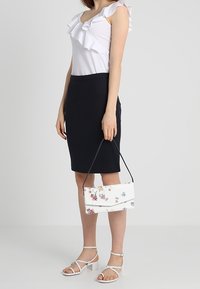 Hvid blomsterhandtaske med en lås, båret sammen med en sort pencilskirt og en hvid volangtop. Model står med strappy hvide sandaler.