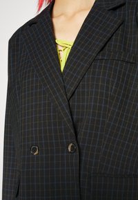 Černobílý kostkovaný dvouřadý blejzr s širokými klopami, na sobě má neonově zelený top s šněrováním, částečně viditelný na hrudníku.