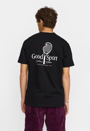 Zwarte katoenen t-shirt met een witte graphic op de achterkant met tekst en een ontwerp van een tennisracket. Model draagt paarse suède broek.