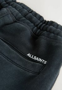 Detail čierneho elastického pásu a vrecka teplákov so štítkom "ALLSAINTS" vytlačeným nad švom vrecka.