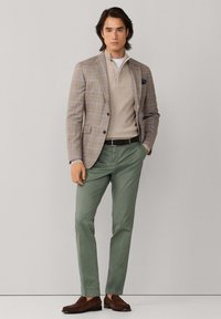 Lichtgrijs geruit blazer met donkere knopen, beige gebreide trui, groene broek en bruine loafers. Soepele texturen en een getailleerde pasvorm.