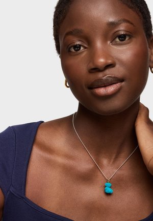 Collana d'argento con un pendente blu a forma di due forme arrotondate sovrapposte, indossata su un collo nudo, su uno sfondo neutro.