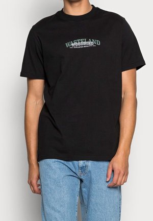 Osoba w czarnej koszulce z napisem "WASTELAND WOODBIRD ISOLATED REALITY" i jasnoniebieskich dżinsach, stojąca na jednolitym tle.