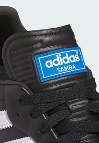 Czarne skórzane trampki o teksturowanym wykończeniu, z białymi paskami, czarnymi sznurowadłami oraz niebieską metką z logo Adidas Samba na języku.