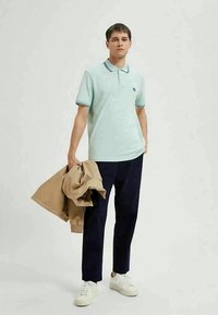 Šviesiai mėlynas polo marškinėliai su apykakle, tamsiai mėlynos kelnės ir balti sportbačiai. Laiko rusvos spalvos švarką; minimalistinis dizainas su tamsiomis akcentinėmis detalėmis.